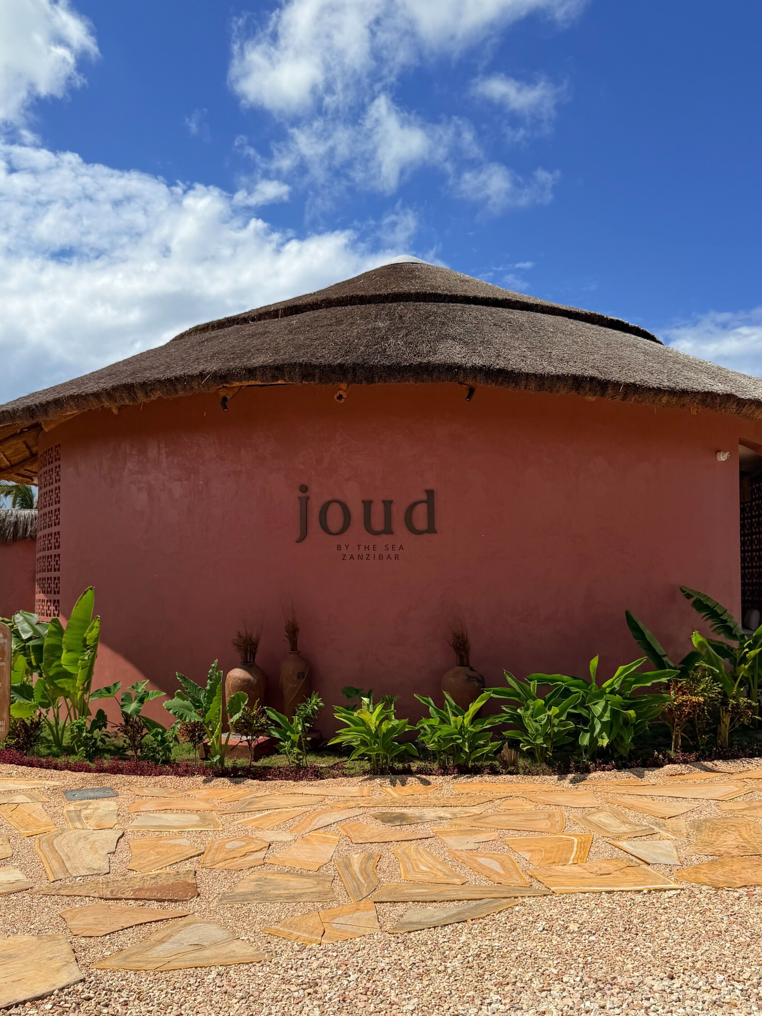 A building with a thatched roof and a wall with the text 'joud by the sea Zanzibar.'