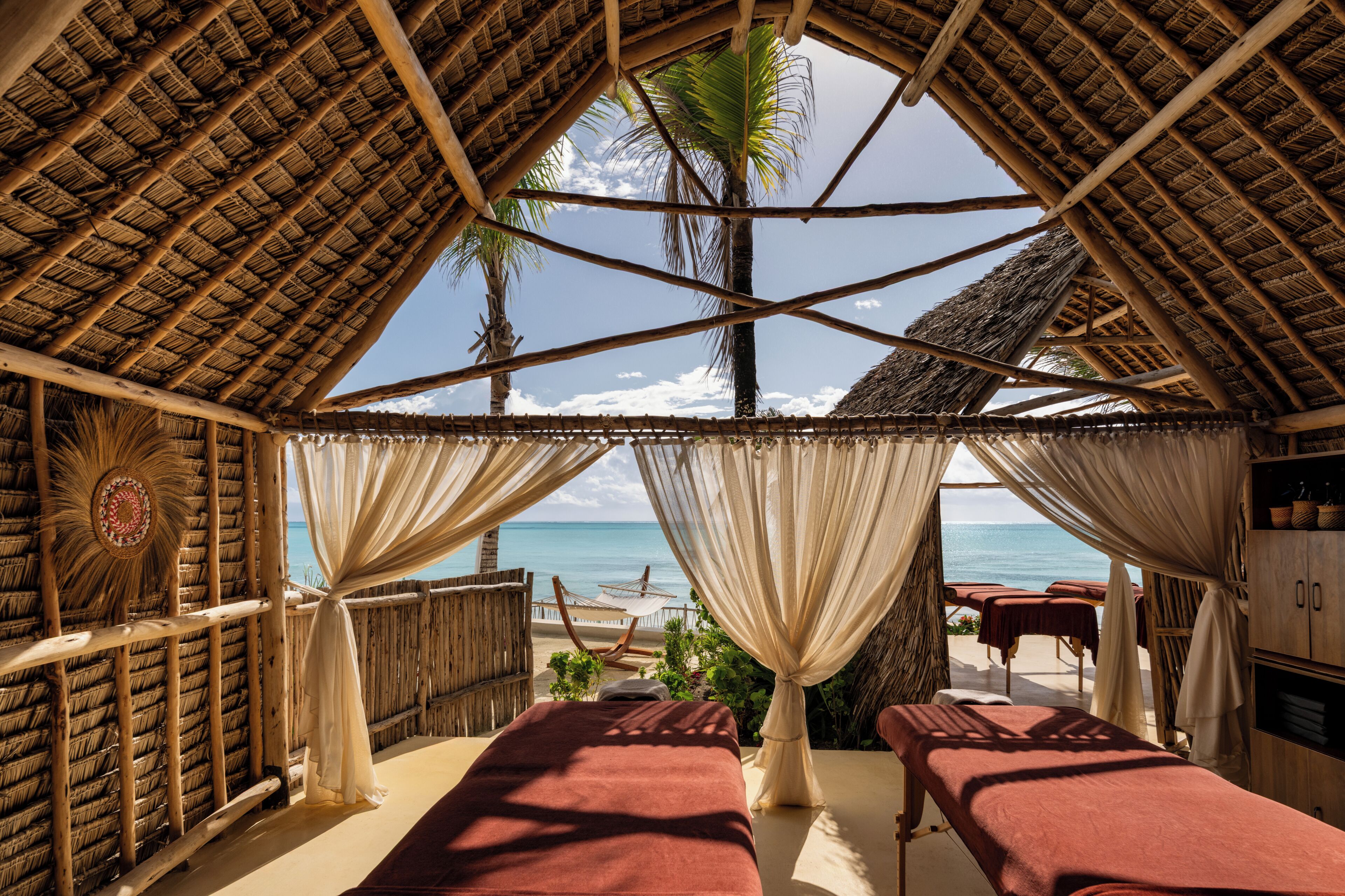 A beachside room with a thatched roof, two massage tables, and a view of the ocean.