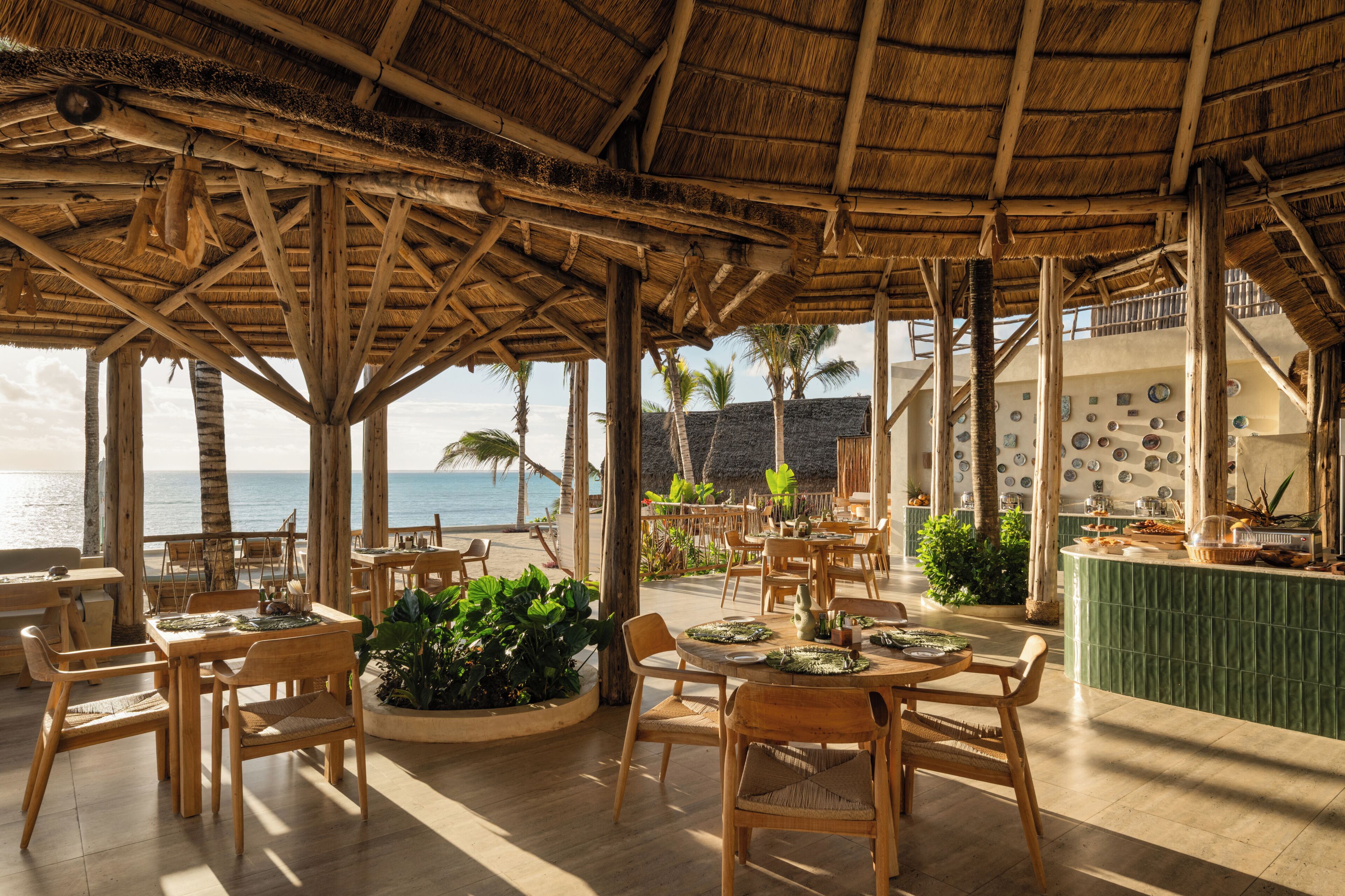 An outdoor dining area with wooden tables and chairs, a thatched roof, and a view of the sea.