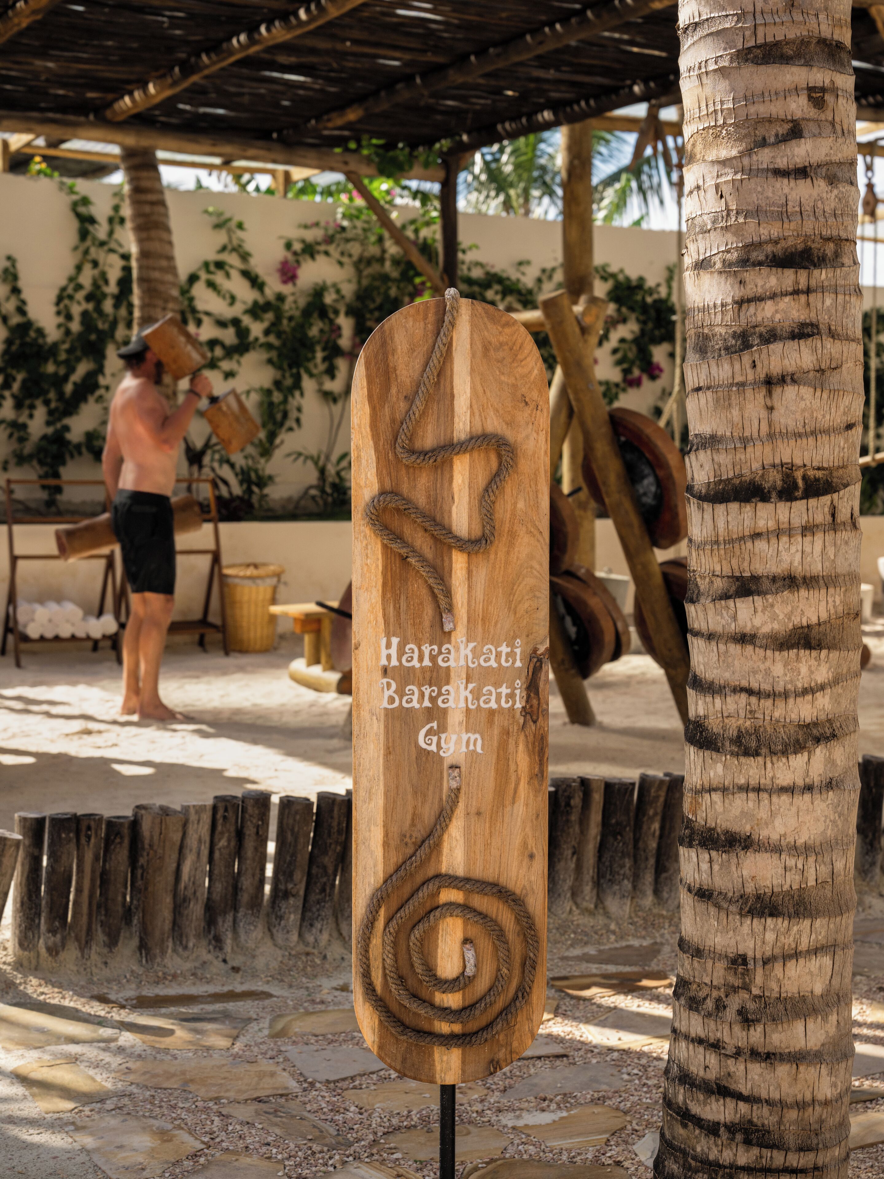 Een houten bord met een touwdesign en de tekst 'Harakati Barakati Gym' in een tropische omgeving met palmbomen en een persoon op de achtergrond.