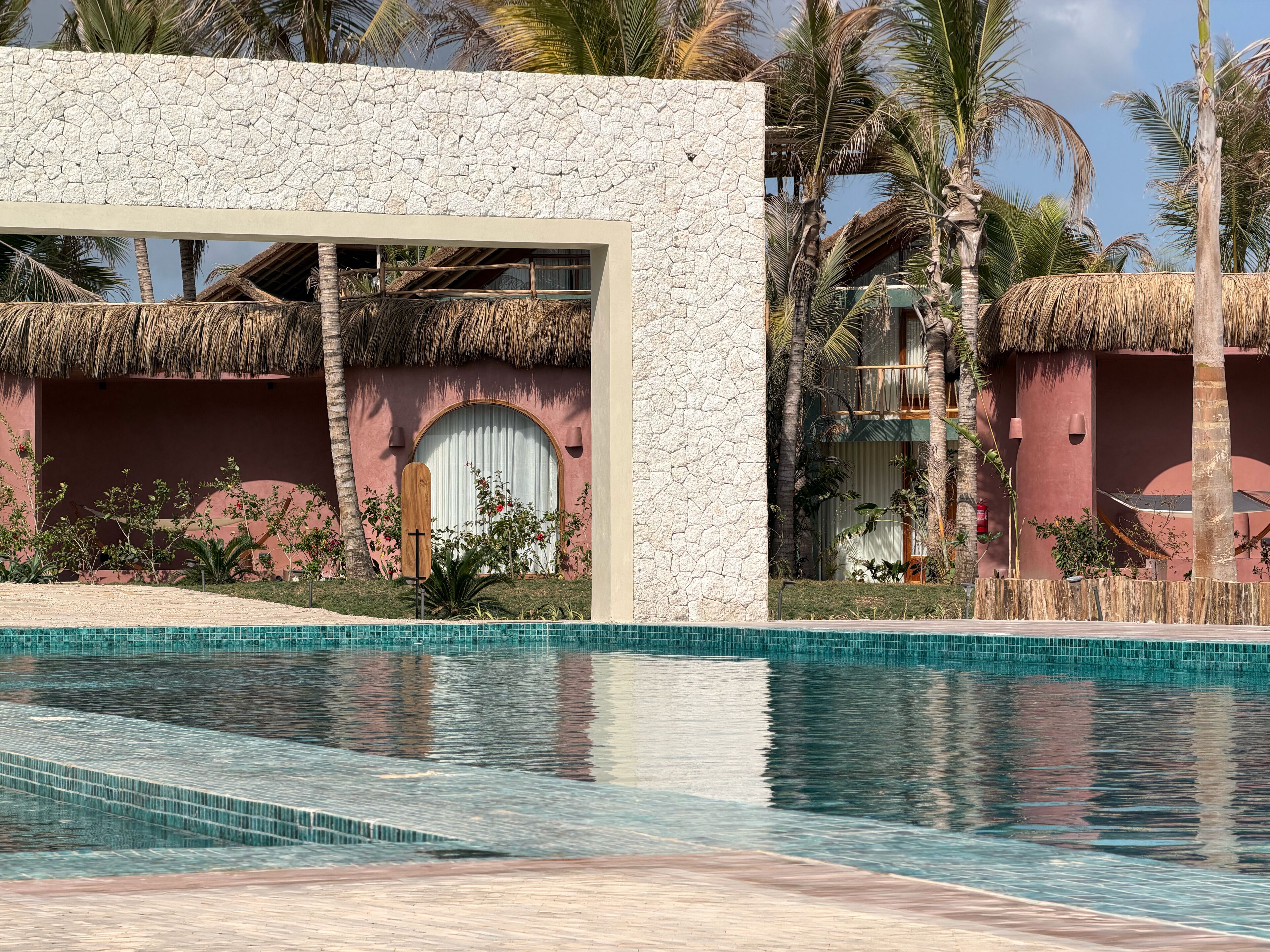 Un área de piscina con una estructura de techo de paja, una pared de piedra y vegetación tropical.