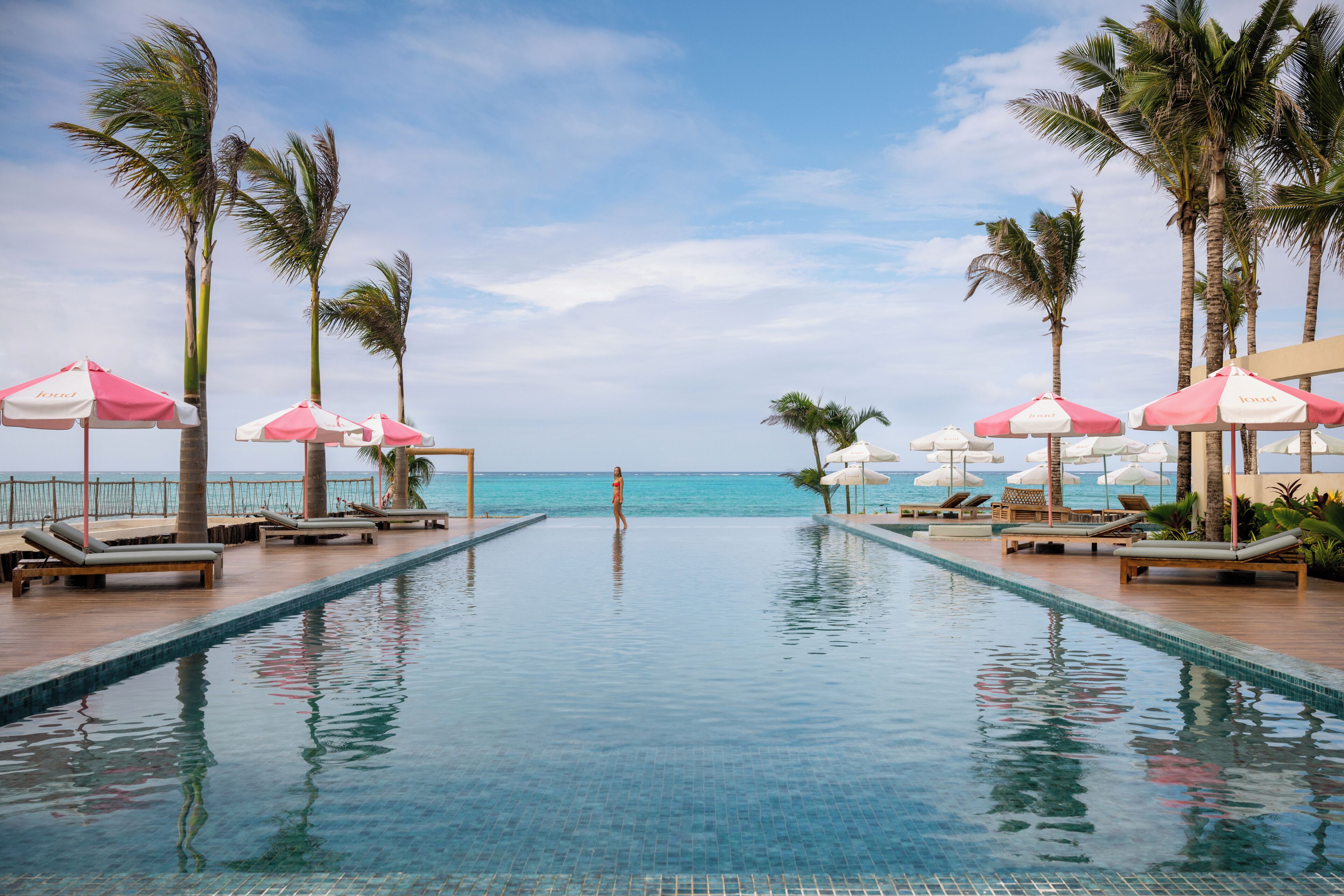 Une piscine avec des chaises longues et des parasols, des palmiers et une vue sur l’océan.