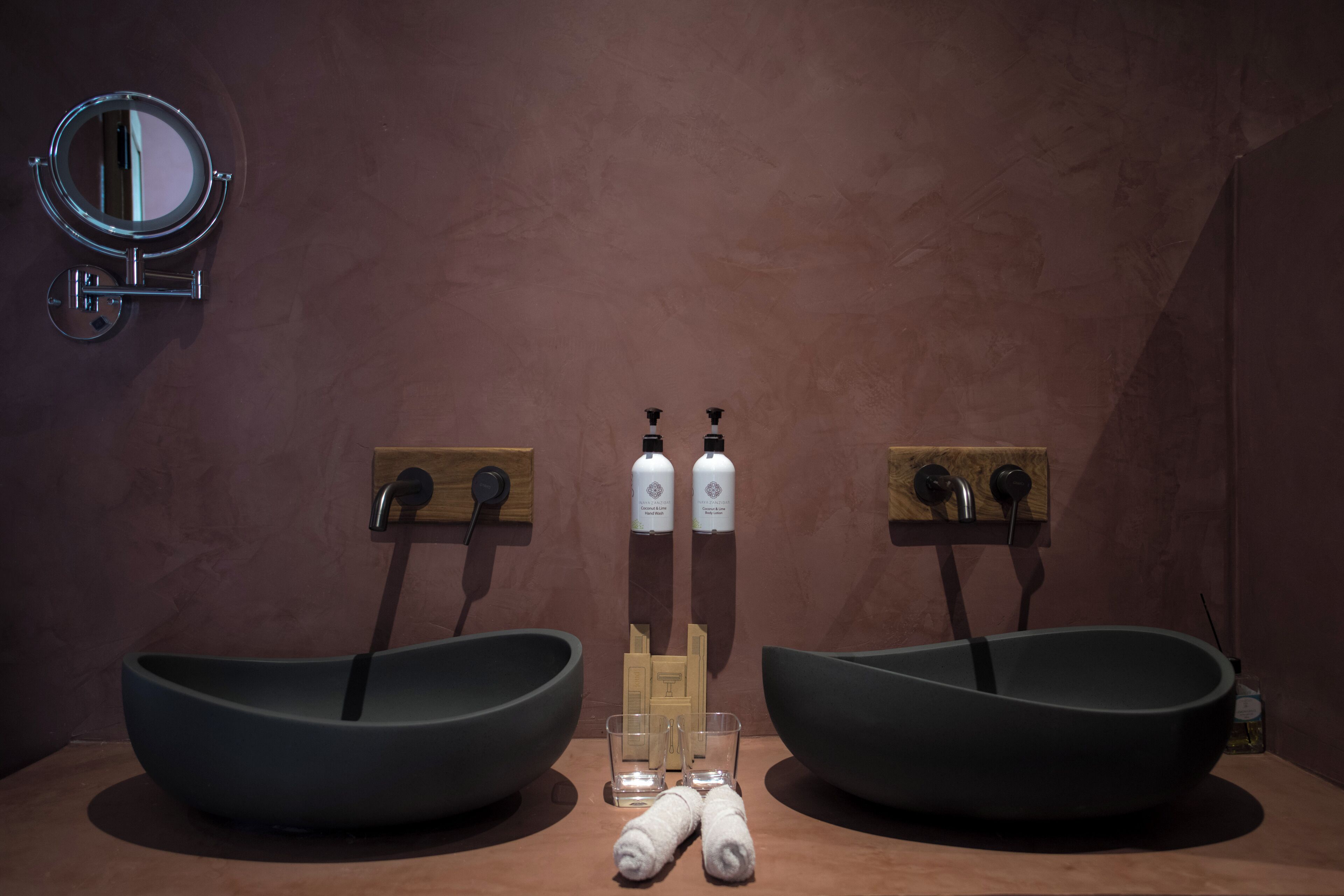 Baño con dos lavamanos grandes y oscuros, un espejo redondo y dos botellas de desinfectante para manos sobre un panel de madera.