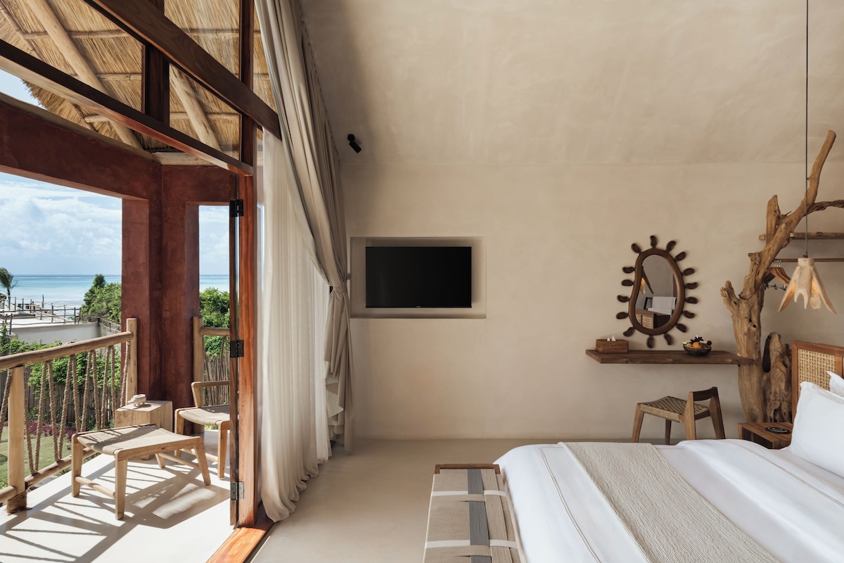 A bedroom with a bed, a TV, a wooden chair, a small table, and a view of the ocean.