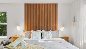 A modern bedroom with a large bed, wooden headboard, bedside lamps, and a view of greenery outside. - Hideaway / Hot Tub / Sunna / Lake / Kayaks / Pool / Game Room / Cold Plunge (Pocono Mountain Lake Estates)