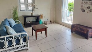 A living room with a sofa, coffee table, and a TV stand. There is a window with blinds, a decorative tree, and a view of greenery outside.