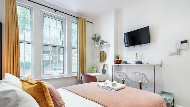 A hotel room with a large bed, a desk with a chair, a TV, and a window with curtains. - Charming Cozy Studio Apartment (London)