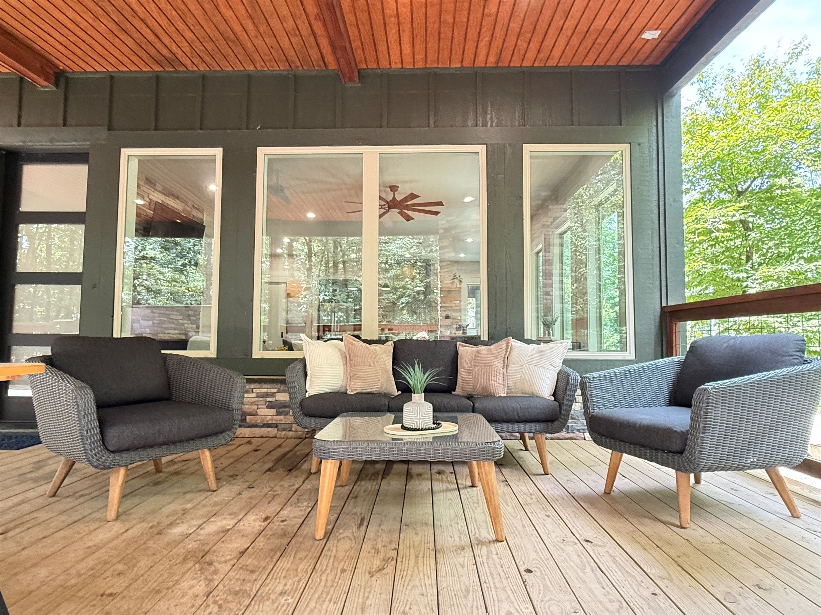 A covered outdoor seating area with wicker furniture, a central coffee table, and large windows offering views of the outdoors.