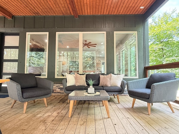A covered outdoor seating area with wicker furniture, a central coffee table, and large windows offering views of the outdoors.