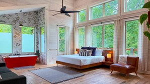 A spacious bedroom with a large bed, a red bathtub, and multiple windows offering a view of greenery. - Ride ATVs • Luxe King Suite •  Modern-Meets-Cozy Retreat  • Prime Location (Broken Bow)