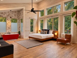 A spacious bedroom with a large bed, a red bathtub, and multiple windows offering a view of the outdoors. - Ride ATVs • Luxe King Suite • EV Charger • Prime Location • Sleeps 10 (Broken Bow)