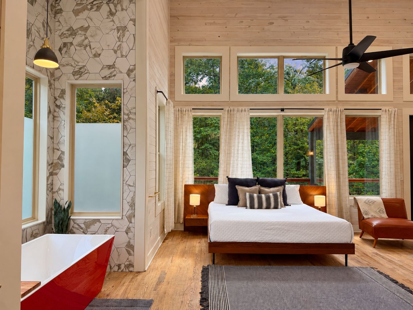 Un dormitorio con una cama grande, cabecera de madera, dos lámparas de noche, una silla roja y vistas a la vegetación a través de amplias ventanas.