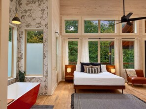 A bedroom with a large bed, wooden headboard, two bedside lamps, a red chair, and a view of greenery through large windows. - Ride ATVs • Luxe King Suite • EV Charger • Prime Location • Sleeps 10 (Broken Bow)