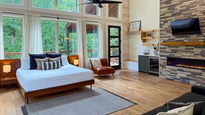 A spacious bedroom with a large bed, a sitting area with a sofa, a fireplace, and a TV mounted on a stone wall. - Ride ATVs • Luxe King Suite •  Modern-Meets-Cozy Retreat  • Prime Location (Broken Bow)