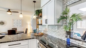 A kitchen with white cabinets, a black marble countertop, and a stainless steel sink. There are ceiling fans, pendant lights, and potted plants.