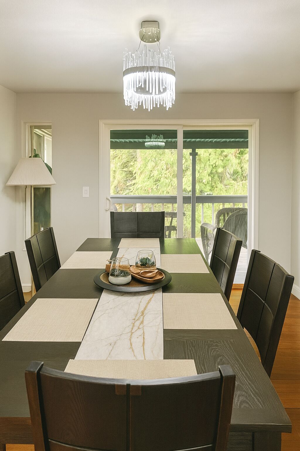 A dining room with a long table, black chairs, a chandelier, and a view of greenery outside.
