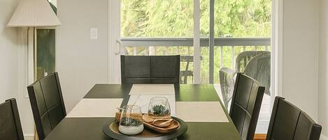 A dining room with a long table, black chairs, a chandelier, and a view of greenery outside.