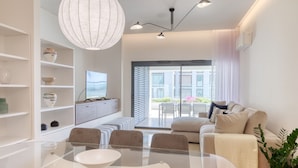 Un salon moderne avec une table à manger en verre, un canapé et un téléviseur posé sur un meuble blanc.