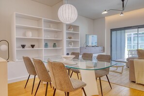 Ein modernes Wohnzimmer mit einem Esstisch mit Glasplatte, beigen Stühlen, einer weißen Regaleinheit und einem großen Fenster mit Jalousien.