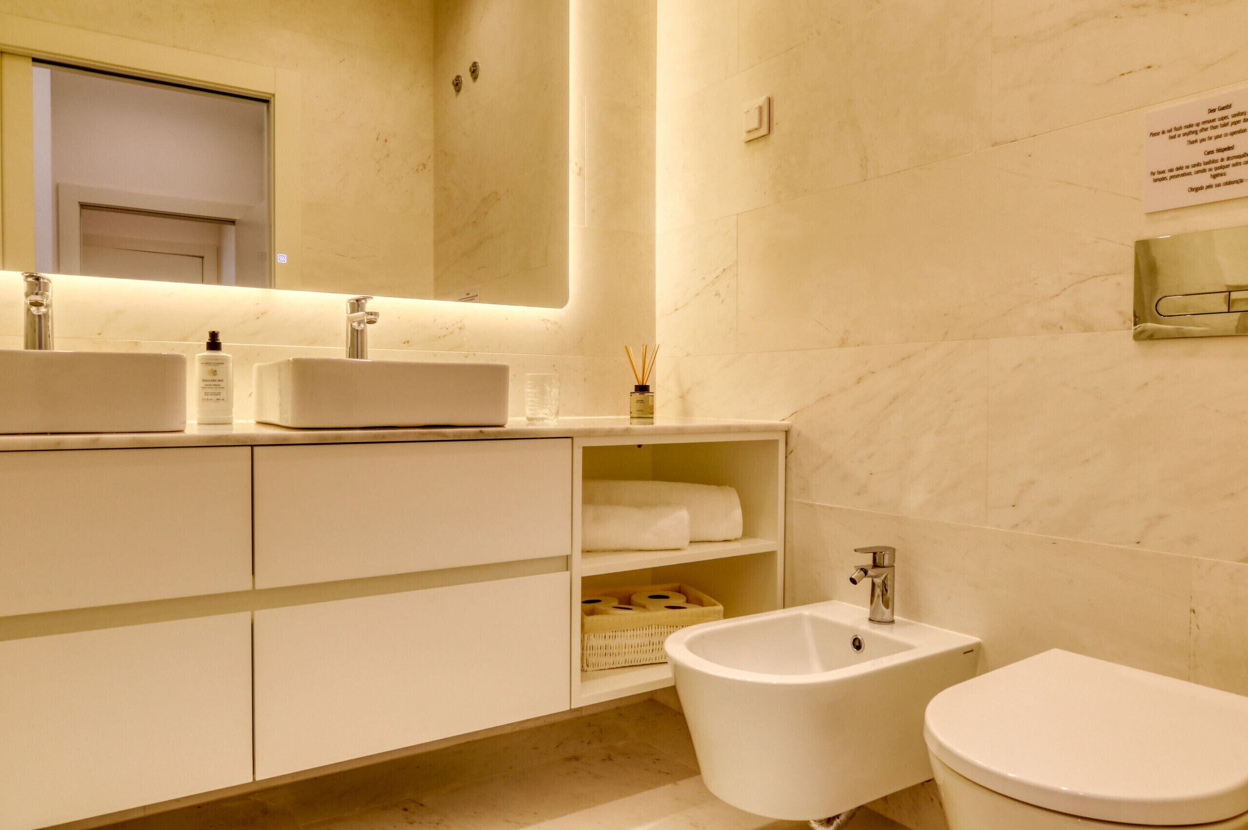 Uma casa de banho moderna com um espelho grande, duas lavatórios, um bidé e uma sanita. Existem prateleiras com toalhas e produtos de higiene pessoal.