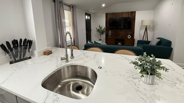 A modern kitchen with a marble countertop, stainless steel sink, and a dining area with a green sofa and a plant.