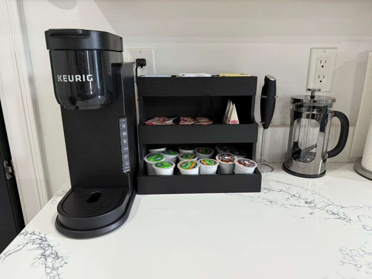 A coffee machine with a coffee pod holder below it, a glass carafe, and a power outlet on the wall.