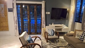 Een woonkamer met een bank, twee stoelen, een glazen salontafel, een televisie en een schuifpui naar een buitenruimte.