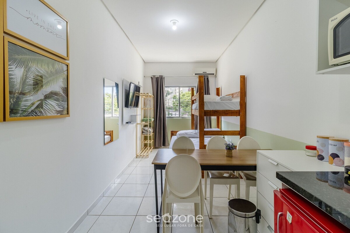 A compact hotel room with a bunk bed, a dining table with chairs, a small kitchen area, and a TV.