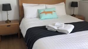 Un letto rifatto con cura, con lenzuola bianche, un cuscino color petrolio con un motivo a volpe e un asciugamano bianco piegato sul letto. Ci sono due comodini con lampade, uno per lato del letto.
