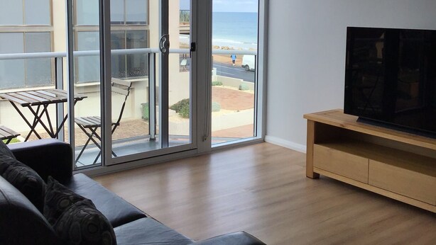 Un soggiorno con un'ampia porta-finestra che dà su un balcone, una TV a schermo piatto su un mobile in legno e un divano scuro.