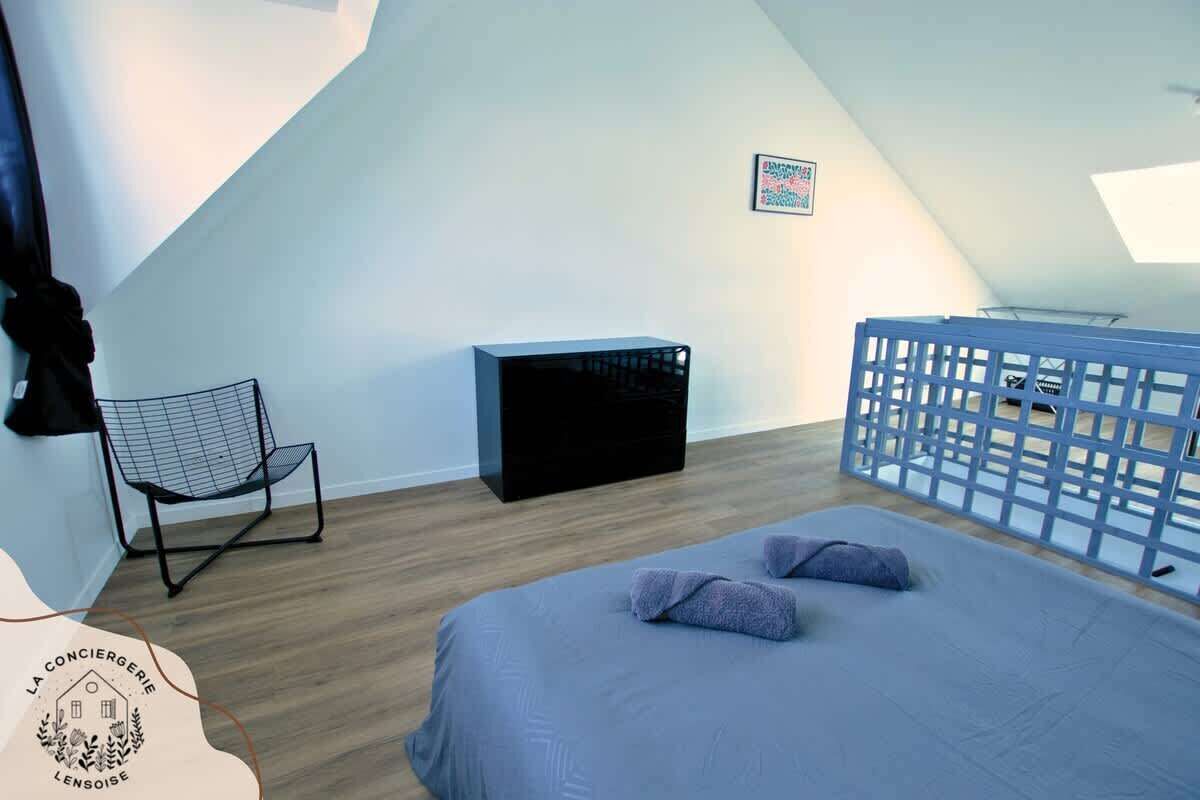 A bedroom with a sloped ceiling, a bed with pillows, a chair, a black storage unit, and a blue railing.