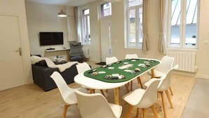 Una habitación con una mesa de póker verde rodeada de sillas blancas, un sofá gris y un televisor en la pared.