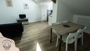 Una habitación moderna y bien iluminada, con una mesa de comedor y sillas, un sofá negro, un televisor pequeño y una kitchenette al fondo.