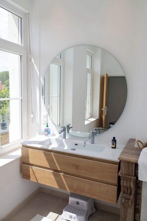 Un bagno con uno specchio tondo grande, un lavabo bianco con due rubinetti e un mobile contenitore in legno con cassetti.