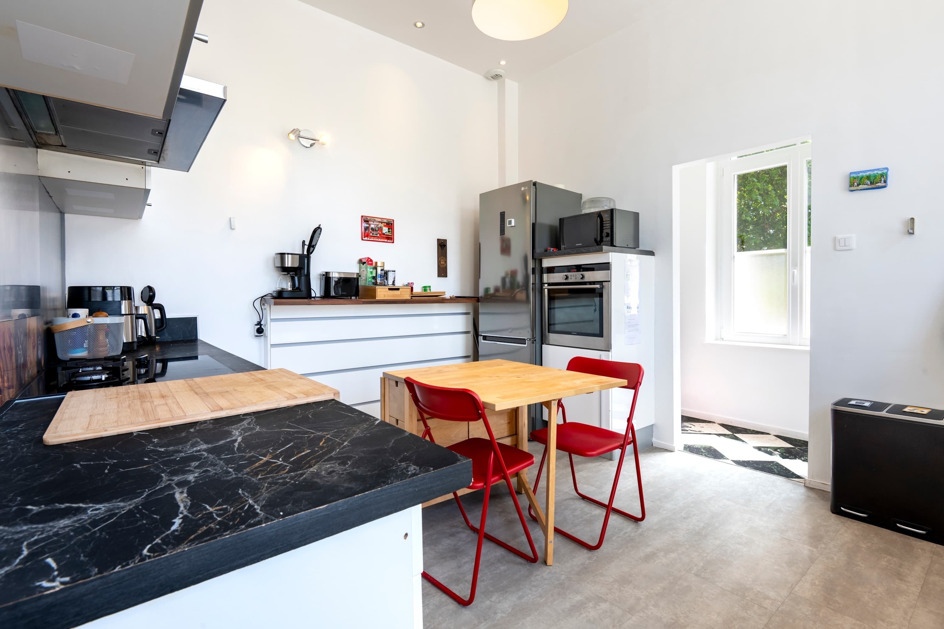 A modern kitchen with a marble countertop, red chairs, a wooden table, and a refrigerator.