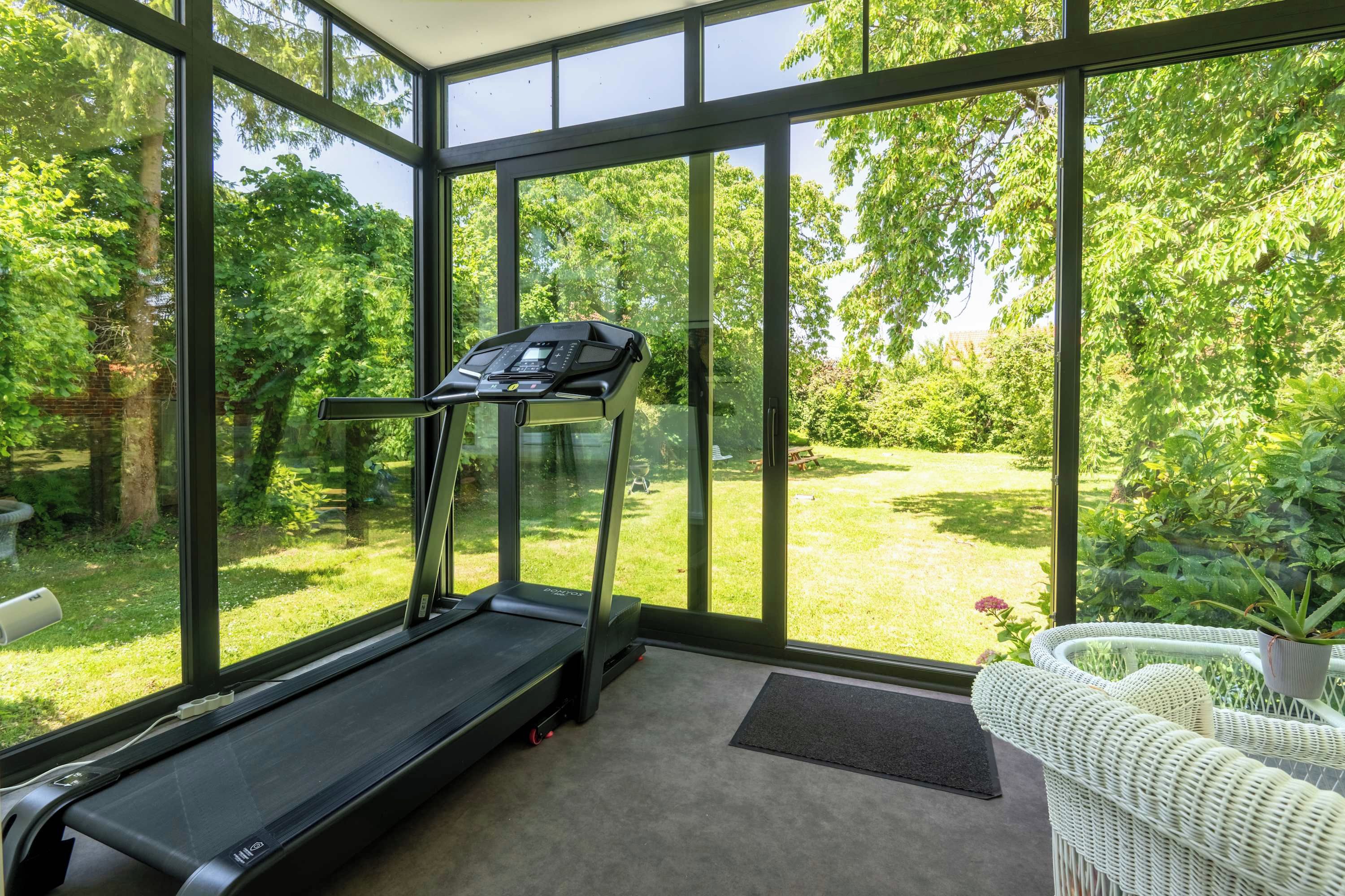Ein Zimmer mit Laufband, großen Fenstern und Blick auf einen Garten.
