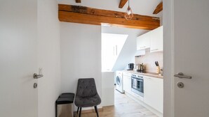 A narrow hallway leading to a kitchen with a washing machine, a chair, and a trash bin.
