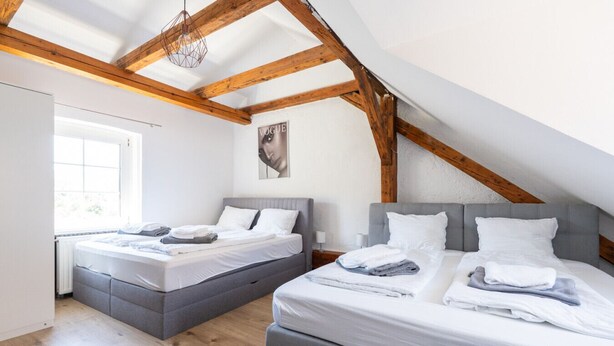 A bedroom with two beds, wooden beams, a large window, and a ceiling light fixture.