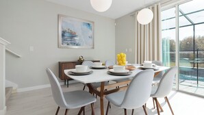 A modern dining area with a white table set for four, grey chairs, and a view of a swimming pool through large windows. - Luxury Townhome near attractions! (Kissimmee)