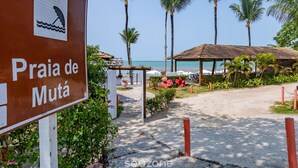 Placa à beira-mar para Praia de Mutá, com um caminho que leva a uma área de praia com palmeiras e um espaço de estar coberto.