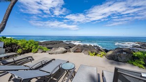 Un área de descanso junto a la playa con sillones y una mesa, con vista al océano, terreno rocoso y cielo despejado.