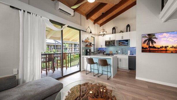 Una cocina moderna con barra, zona de comedor con mesa de vidrio y balcón con mobiliario exterior.