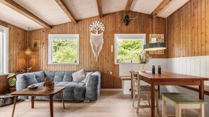 A cozy room with wooden walls, a grey sofa, a wooden table, and two windows with views of greenery. - Amazing home in Melby with sauna (Melby)