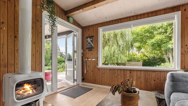 A cozy room with a wood stove, a large window with a view of trees, and a door leading outside. - Amazing home in Melby with sauna (Melby)