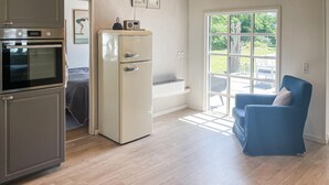 Une cuisine compacte équipée d’un réfrigérateur, d’un four et d’un four à micro-ondes. Une chaise bleue est placée près d’une grande fenêtre donnant sur l’extérieur.