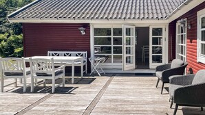 Une terrasse en bois avec une table et des chaises, un bâtiment rouge avec un toit en tuiles et une porte vitrée donnant sur l’intérieur.