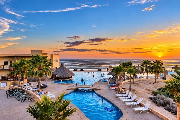 A resort pool area with a view of the ocean at sunset, including a swimming pool, lounge chairs, and palm trees.