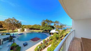 A pool area with a curved design, surrounded by greenery and a clear view of the ocean. - Gemini Resort - Unit 19 (Golden Beach)