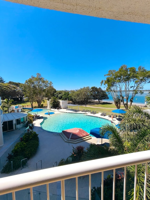 A swimming pool with a curved design, surrounded by lounge chairs and umbrellas, with a view of the sea and trees in the background.