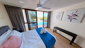 A hotel room with a bed, a bench, a ceiling fan, a window with curtains, and artwork on the wall. - Gemini Resort - Unit 19 (Golden Beach)